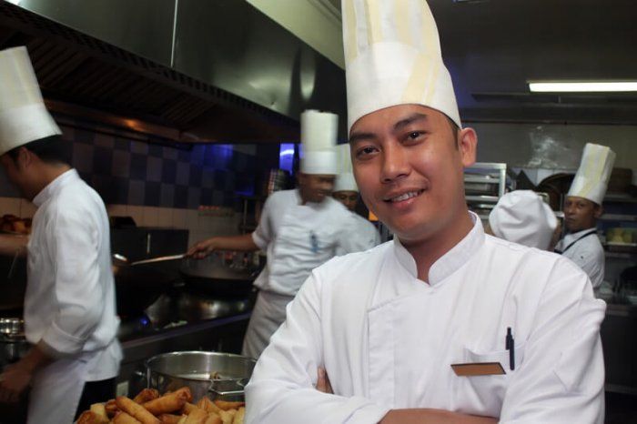 asian chef standing in a kitchen with several assistant chefs in the background doing work