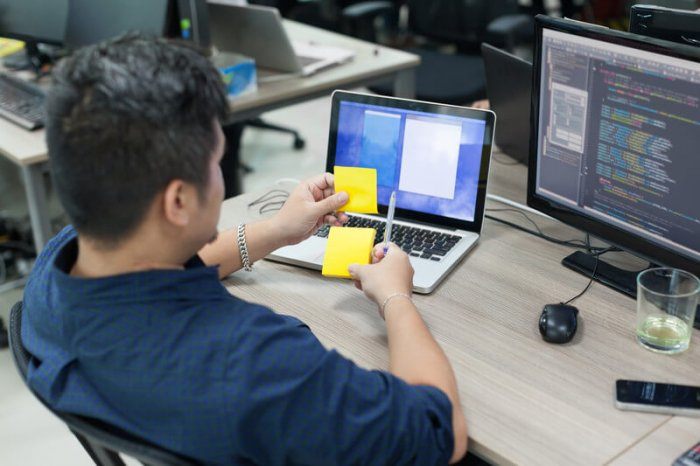 a man with dark hair sitting at an office desk doing we development and looking at two sticky notes in his hand