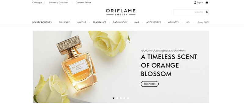 Oriflame website screenshot showing a white tabletop with two roses and a bottle of perfume. 