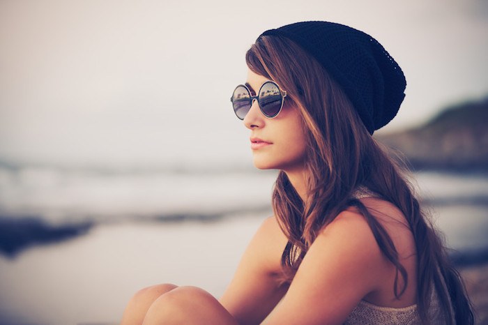 young woman sitting on the beach with vintage fashion sunglasses and a black beanie hat