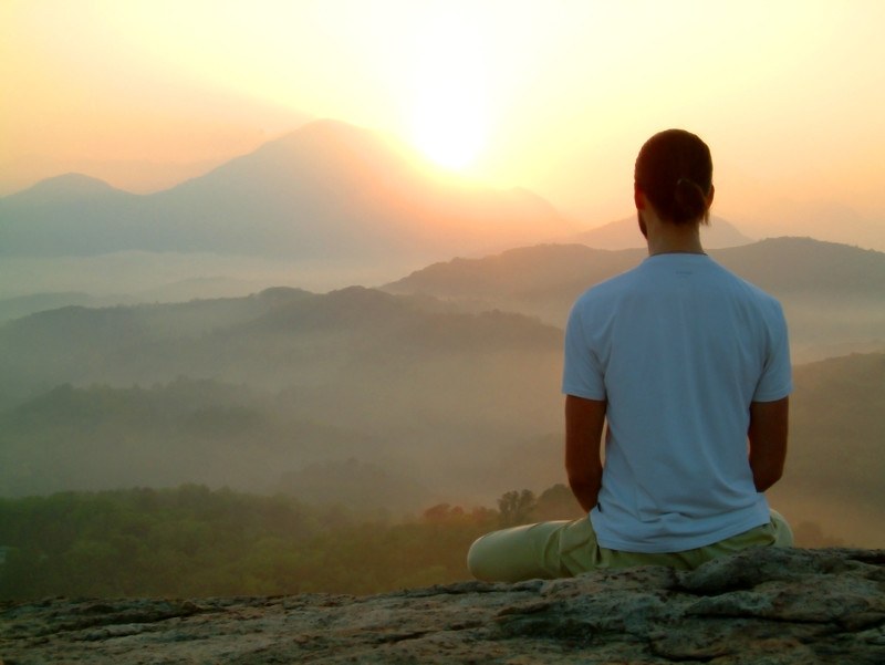 young man sitting on a cliff's edge watching the sun rise over misty mountains while meditating