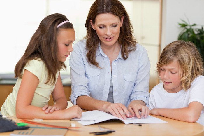 middle aged woman homeschooling two young girls with paper and pen at a desk in their home