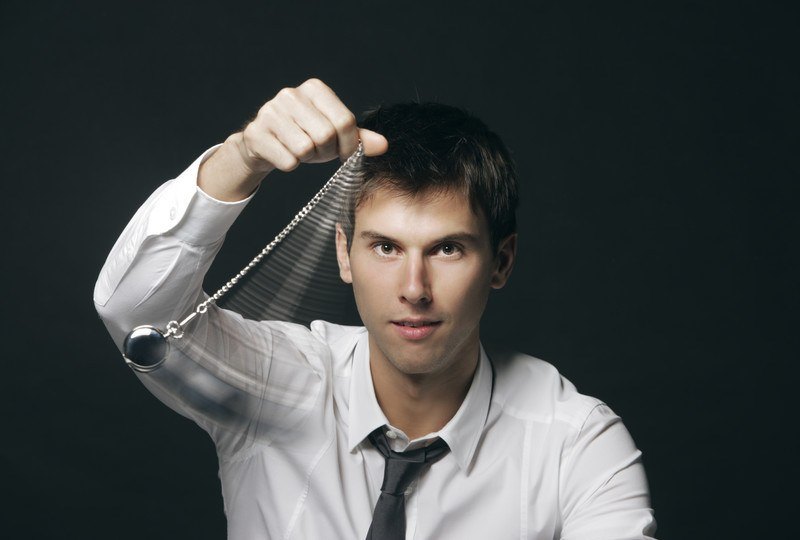 A man in a white shirt with a tie sits in front of a dark background as he swings a watch on a chain, representing the best affiliate programs for hypnosis.