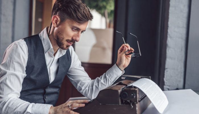 A man in a white suit and business vest sits at a table and looks at a paper that he's typing on a vintage typewriter, representing the best screenwriting affiliate programs.