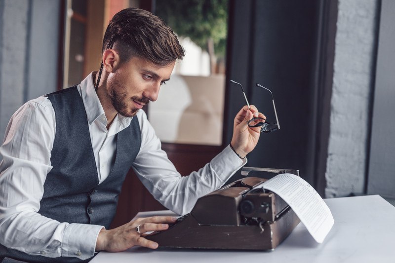 A man in a white suit and business vest sits at a table and looks at a paper that he's typing on a vintage typewriter, representing the best screenwriting affiliate programs.