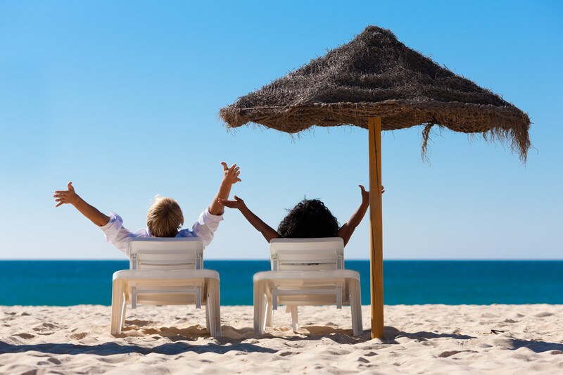 This photo shows a man and a woman in lounge chairs under an umbrella on a beach, facing the ocean with their arms open above their heads in a pose of victory and freedom, representing the best vacation affiliate programs.