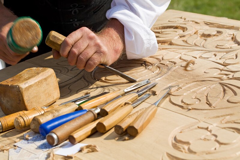 This photo shows the hands of a wood craftsman working with a chisel and mallet on a relief carving, with a set of tools lying at the edge of the wood project, representing the best woodworking affiliate programs.