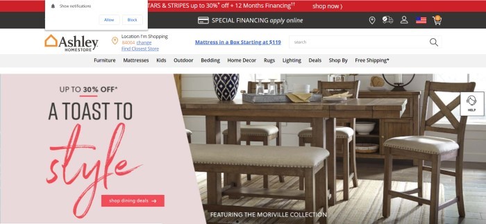 This screenshot of the home page for Ashley Furniture shows a dark wooden dining set with beige cushions on the right side of the screen and a pink advertisement for 30% off on the left side of the page.