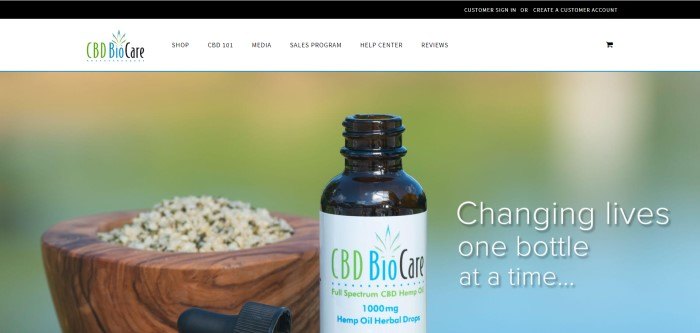 This screenshot of the home page for CBD BioCare shows an open dropper bottle of a CBD BioCare product near a wooden bowl filled with ground herbs, and the phrase 'Changing Lives One Bottle At A Time' in white text on a multi-colored, blurred background.