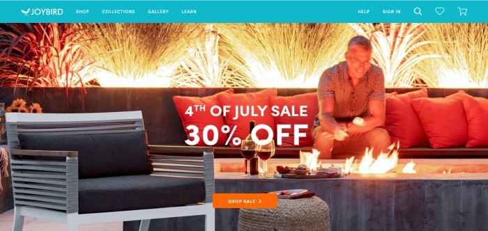 This screenshot of the home page for Joybird shows a man sitting on some outdoor patio furniture, toasting a marshmallow over a fire, while white fireworks explode in the background. 