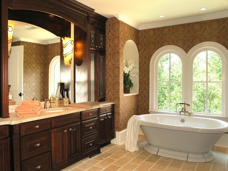 This photo of a bathroom shows a sink with dark wooden cabinets, a tan tiled floor, and a large white bathtub with a white bath towel on its edge, near a set of arched windows with greenery beyond them, representing the best kitchen and bath affiliate programs. 