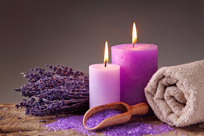 This photo shows two purple candles, a rolled up beige colored towel, a scoop of light purple bath salts and a bouquet of dried lavender on a table together, representing the best candle affiliate programs.