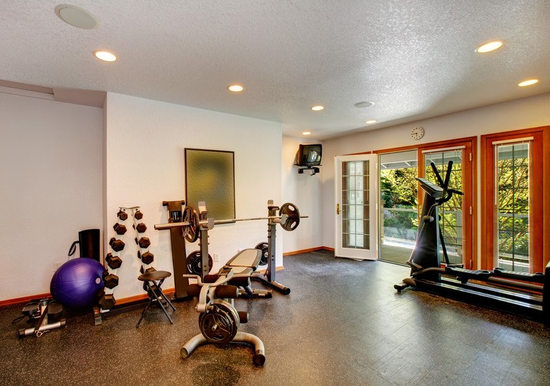 This photograph shows a room with a large window, white walls, and a brown floor with several pieces of exercise equipment in it, including a treadmill, weight racks with weights, and a weight bench, representing the best home gym affiliate programs.