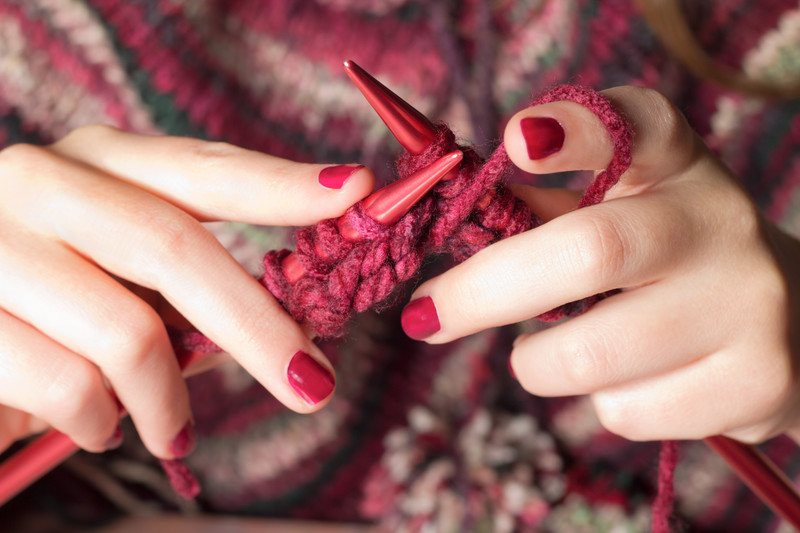 This photo shows a woman's hands with red painted fingernails, holding red knitting needles and knitting something in a red, brown, and white variegated yarn, representing the best knitting affiliate programs.