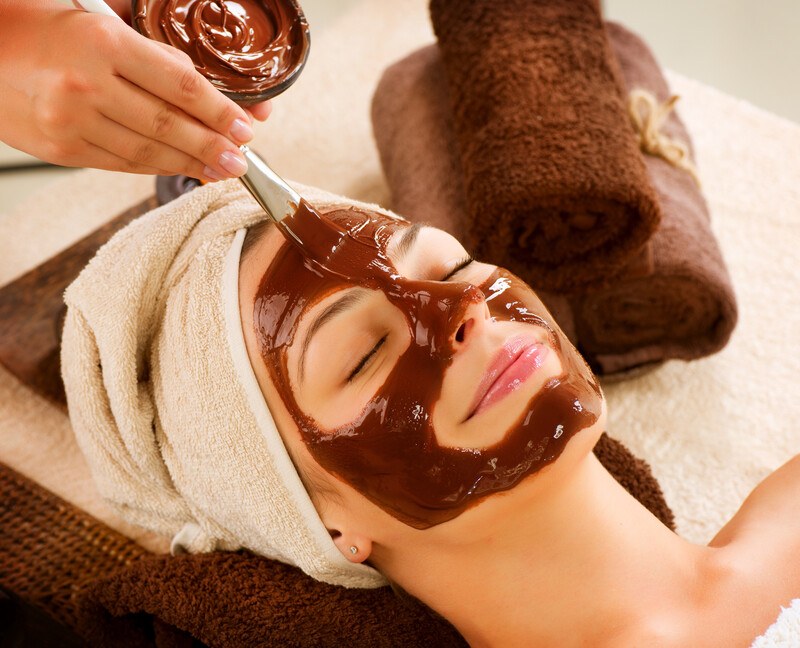 A woman with a towel over her head reclines with her eyes closed while someone paints a chocolate mask onto her face, representing the best skin care affiliate programs.