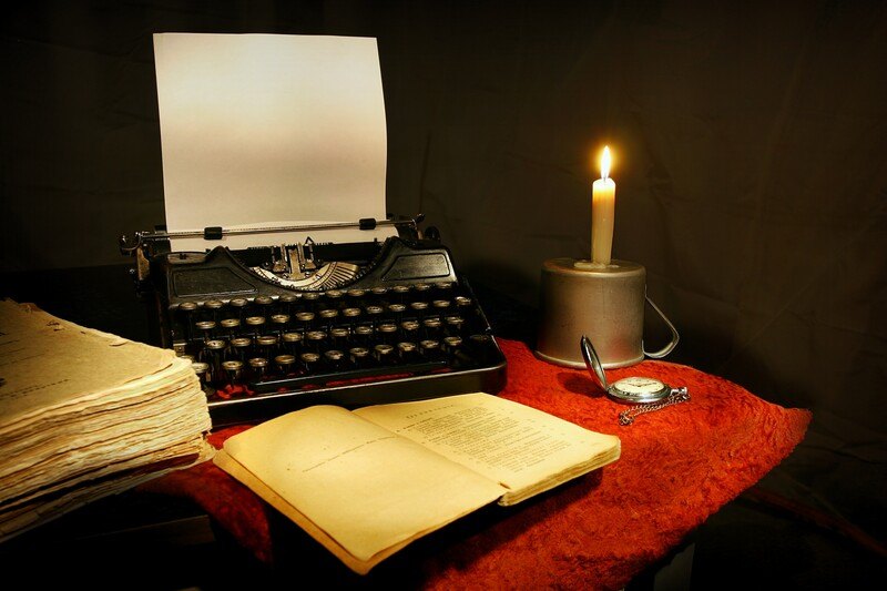 This photo shows an old-fashioned typewriter with paper in it, an open book, a red cloth and a lit candle on a table in a dark room, representing the best writing affiliate programs.