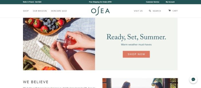 This screenshot of the homepage for OSEA has a white background, with a beige insert on the right side that includes some summer-geared text and a shopping link and a photo on the left side showing a woman's hands as she holds an OSEA product on a picnic blanket that also contains another OSEA product, a wooden tray of vegies, and a wooden tray of strawberries and blueberries. 