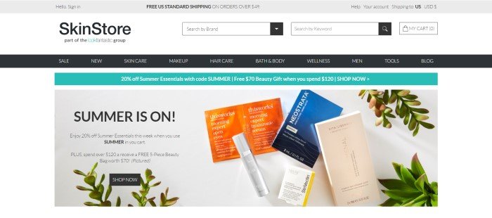 This screenshot of the home page for SkinStore has a white background, a black navigation bar, and an advertisement for a summer sale along with some skin care products in a silver tube, a blue box, an ivory box, and an orange foil packet with sprigs of plants against a gray background.