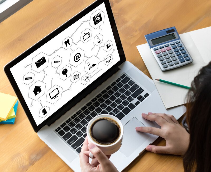 This photo shows the right hand of an unseen person orking on a laptop that has a screen of icons that are connected as if they are automated, while the other hand holds a cup of coffee, representing the best autoresponder affiliate programs.