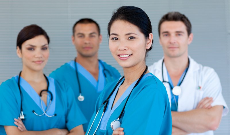 This photo shows two male and two female doctors standing with arms folded and pleasant expressions on their faces in front of a set of gray blinds, representing the best healthcare affiliate programs.