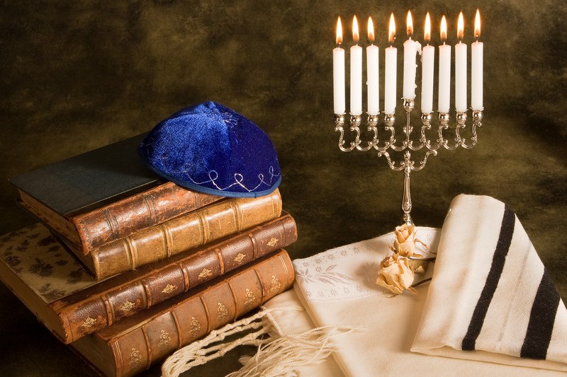 This photo has a blue yarmulka on top of a stack of four brown books, near a lit Menorah on a white cloth, representing the best Hebrew affiliate programs.