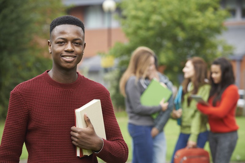 This photo shows a smiling young man in a red sweater, holding a book, while behind him three girls talk together in front of what appears to be a school building, representing the best school affiliate programs.