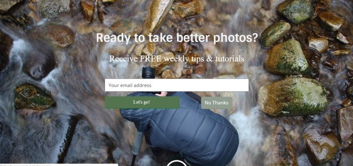 This screenshot of the home page for Digital Photography School includes a large overhead-shot photograph of a person in a black hooded jacket with a nice camera taking a picture of a rocky stream bed, along with white text reading 'Ready to take better photos?' and an opt-in bar.