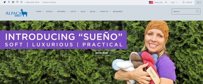  This screenshot of the home page of Alpaca Direct shows a woman in a tan beanie standing near a green hedge with her arms full of various colors of Sueno alpaca yarn.