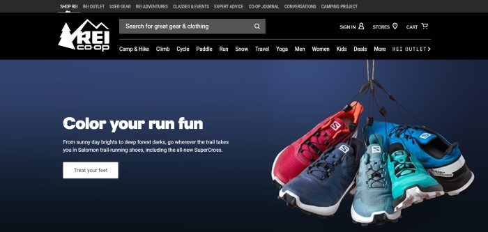  This screenshot of the home page for REI has a black header and navigation bar with white lettering and a white REI logo in the left upper corner, and below it, a page with a dark blue background has a photo of several sneakers hanging by their shoelaces and text in white lettering that reads 'Color your run fun' with an advertisement for running shoes and a white call-to-action button.