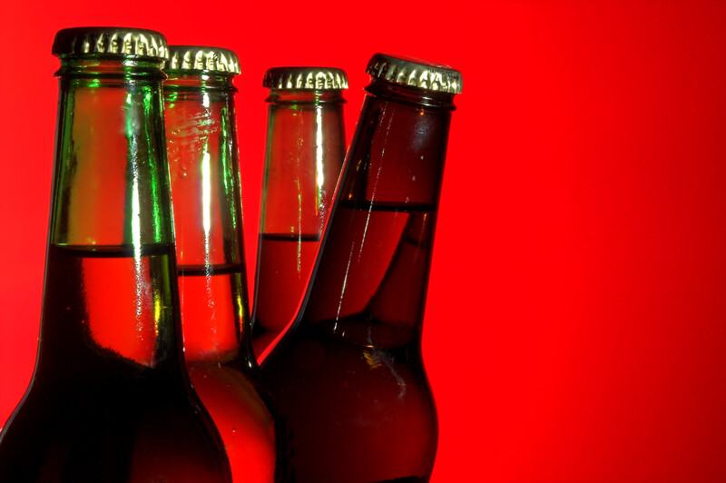 This image has a row of four green-glass beer bottles in front of a red background, with the third bottle leaning forward as if it's peeking out from behind the two bottles in front of it, representing the best home brewing affiliate programs.