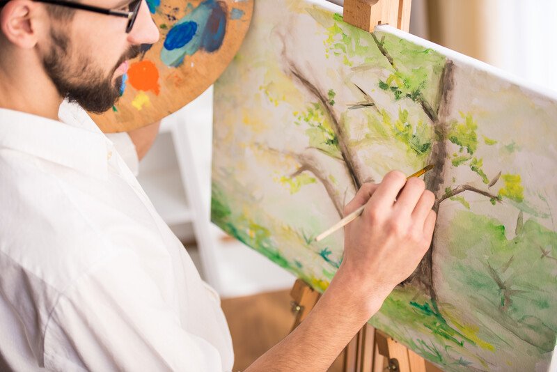 This photo shows a man with dark hair and a beard holding a palette and a paint brush, as if he's about to add more to a painting of some trees on an easel in front of him, representing the best painting affiliate programs.