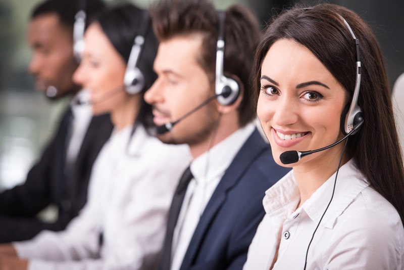 Two men and two women in business clothing and headphones sit at a table with a row of computers, and the woman on the end is looking up and smiling, representing the best telemarketing affiliate programs.