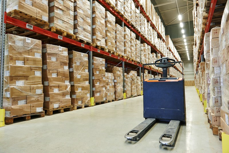 A blue forklift rests in the middle of an aisle between a set of three-story shelves filled with pallets of cardboard boxes, representing the best wholesale affiliate programs. 