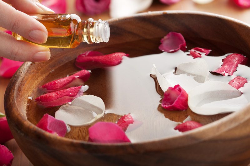 woman putting essential oils into wooden bowl with water and rose petals