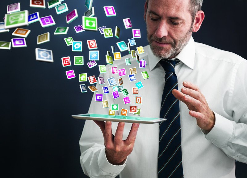 This photo shows a bearded man in business clothes holding a tablet in his hands, while icons for several apps fly in the air around him as if they're emerging from his tablet, representing the best apps affiliate programs.