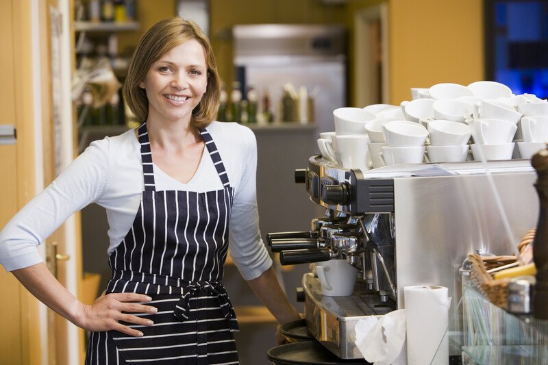 This photo shows a smiling blonde woman in a white shirt and a black and white apron standing in front of an espresso machine and stacks of white espresso cups, representing the best espresso machine affiliate programs.