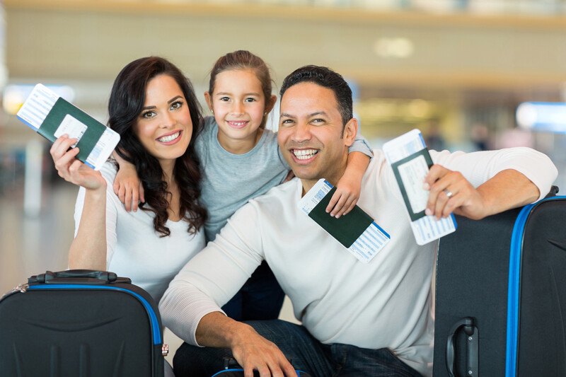  This photo shows a smiling dark-haired woman in a white shirt, a smiling dark-haired man, and a young girl holding passports and boarding passes in the waiting area of an airport, representing the best travelers insurance affiliate programs. 