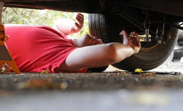 A young man in a red shirt underneath his car repairing it