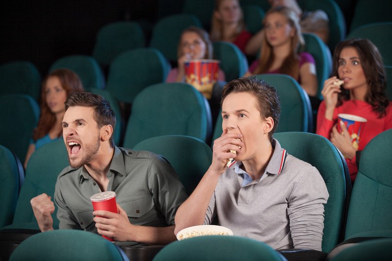 Two men sitting in a theatre watching a movie, with various people behind them