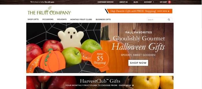 This screenshot of the home page of The Fruit Company has a white background with green and orange elements and black text in the navigation bar, above a photo of a black box with a white spiderweb pattern, which contains green and red apples with white ghost cookies and orange pumpkin cookies, as well as another photo of a pumpkin sitting in front of a rustic wooden wall.