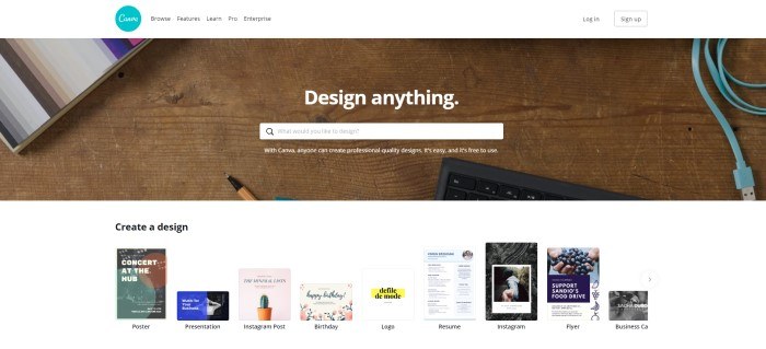 This screenshot of the home page for Canva shows a wooden desk top with the edge of a black keyboard, the edge of a stack of multi-colored paper, and the tip of a pen, along with white text reading "Design anything" and a search bar for getting started.