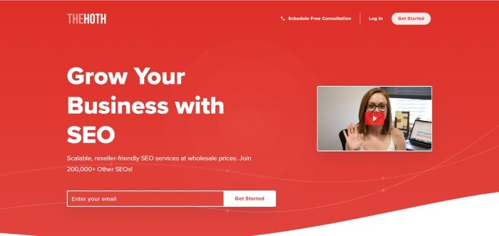 This screenshot of home page for The Hoth has a red background, white lettering inviting customers to grow their business with SEO , and an insert of a video clip with a smiling woman waving at the camera.