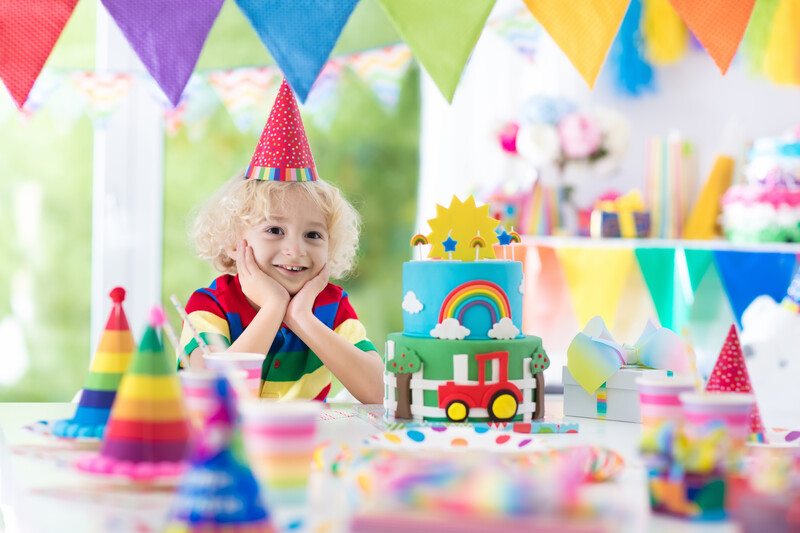 This image shows a blonde child in a red party hat, sitting at the end of a table with his chin in his hands, surrounded by multicolored banners, toys, gift bags, and other party hats that designate each place setting, representing the best party affiliate programs. 