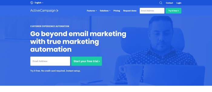 This screenshot of the home page for Active Campaign has a royal blue background with a blue filtered photo of a man in a polka-dot shirt sitting in an office chair, looking at a laptop near a white coffee up, along with white wording that reads "Go beyond email marketing with true marketing automation" and a two green call-to-action buttons.