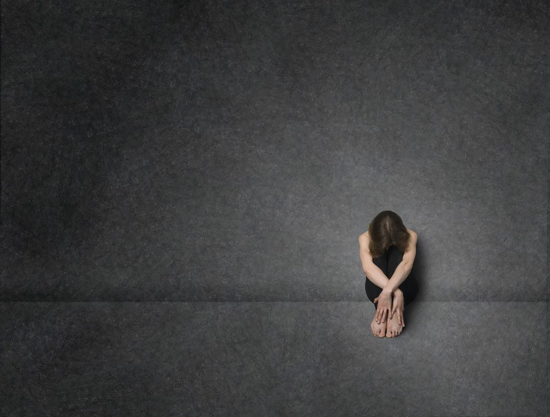 This image shows a single woman with dark hair and black clothing, sitting on the floor in a dark room with gray walls and a gray floor, with her knees bent in front of her, her head on her knees so no one can see her face, and her arms folded protectively over her lower legs, representing the best depression affiliate programs.