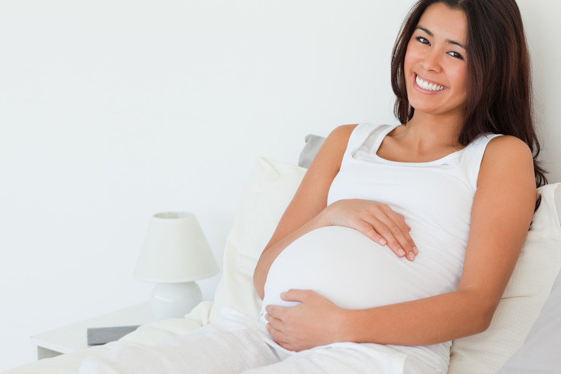 This photo shows a smiling dark-haired woman wearing all white, lying on a white bed in a white room, with both her arms wrapped around her very pregnant belly, representing the best pregnancy affiliate programs.