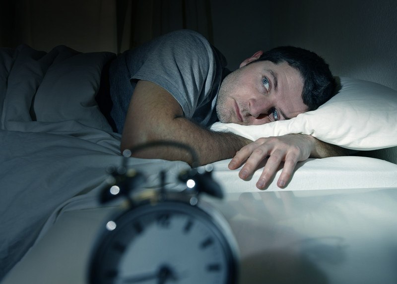 This image shows a man in a gray shirt, lying in bed with an unhappy expression on his face and wide eyes, as he looks toward the alarm clock on the nightstand next to him, representing the best sleep affiliate programs.