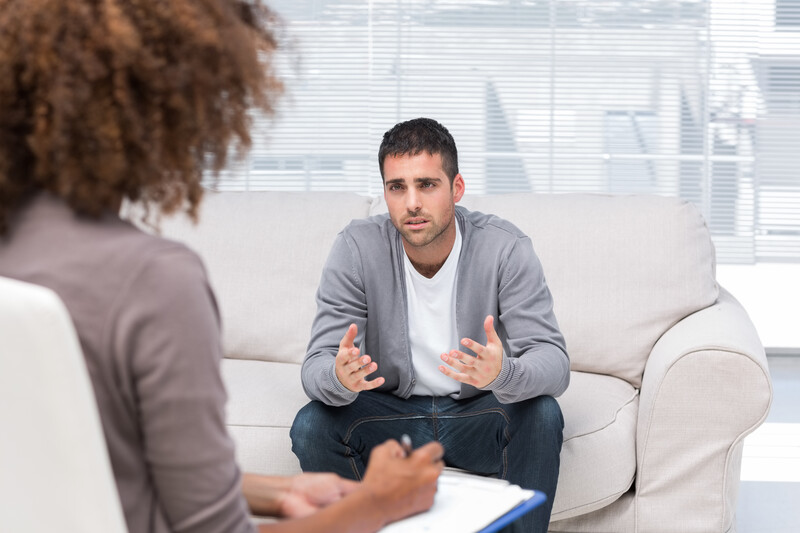 This image shows a dark-haired young man in a white tee shirt, jeans, and an open button-down shirt, who is sitting on a white coach beside a window showing a cityscape beyond it, explaining something to a curly-haired woman facing him with a clipboard on her lap, representing the best therapy affiliate programs.