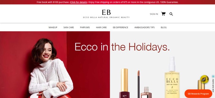 This screenshot of the home page for Ecco Bella has a red header announcing a sale, a white navigation bar, a photo of a smiling brown-haired woman with perfect makeup and a white sweater in front of a red background on the left side of the page, and a photo of Ecco Bella products against a white background on the right side of the page. 