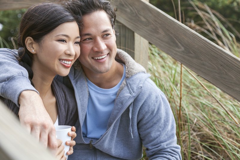 This image shows a smiling Asian woman and men sitting near a wooden fence with grass behind it, holding white cups of a hot drink, representing the best Asian dating affiliate programs.